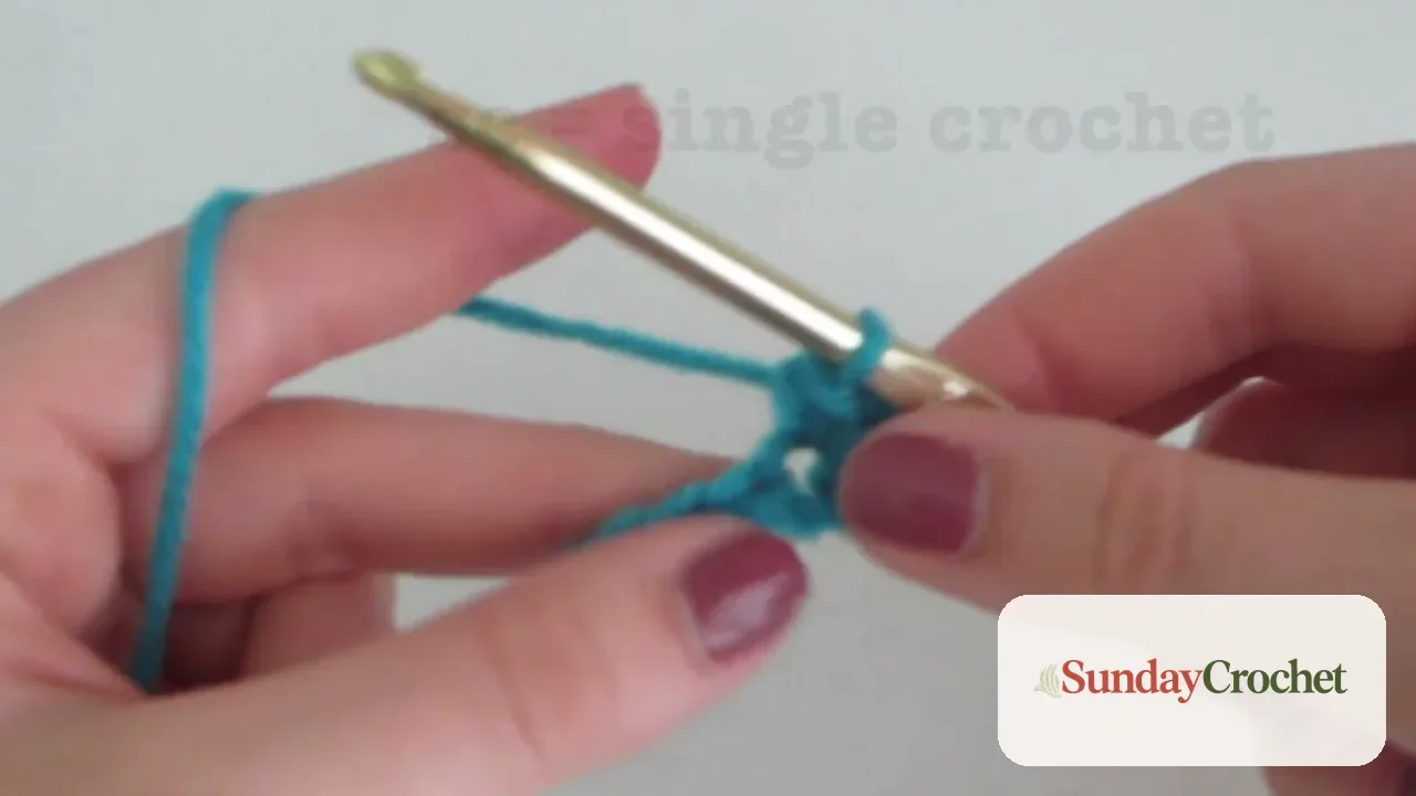 Hands holding blue yarn and a hook while forming a half double crochet stitch
