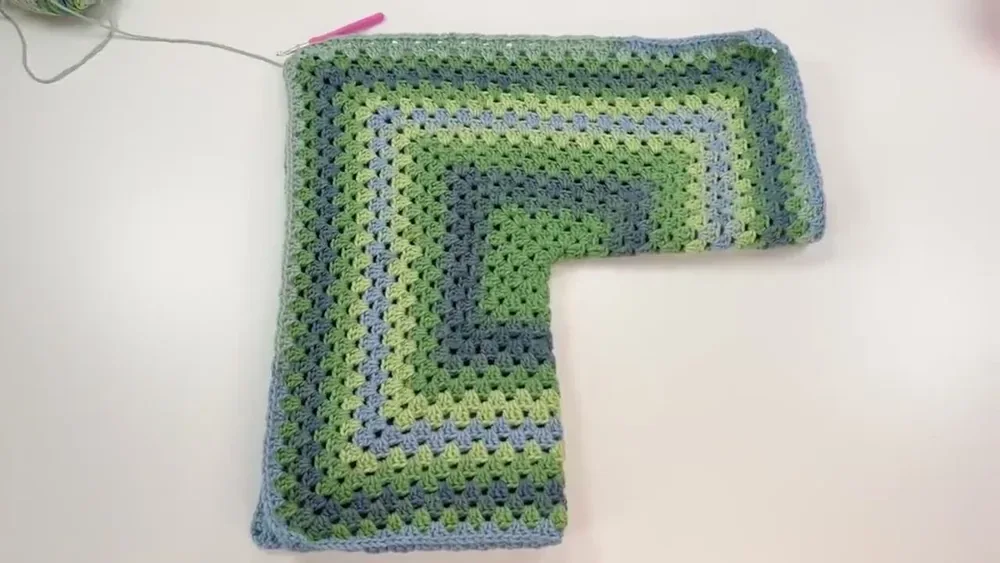 The folded green and blue granny hexagon panel is laid out to check its short side against shoulder width