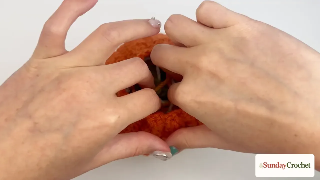 Hands add yarn-scrap filling inside the orange crochet pumpkin before the top is closed