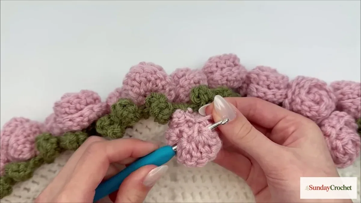 A pink crochet rose is joined with a hook to the green leaf edging around the ivory blanket