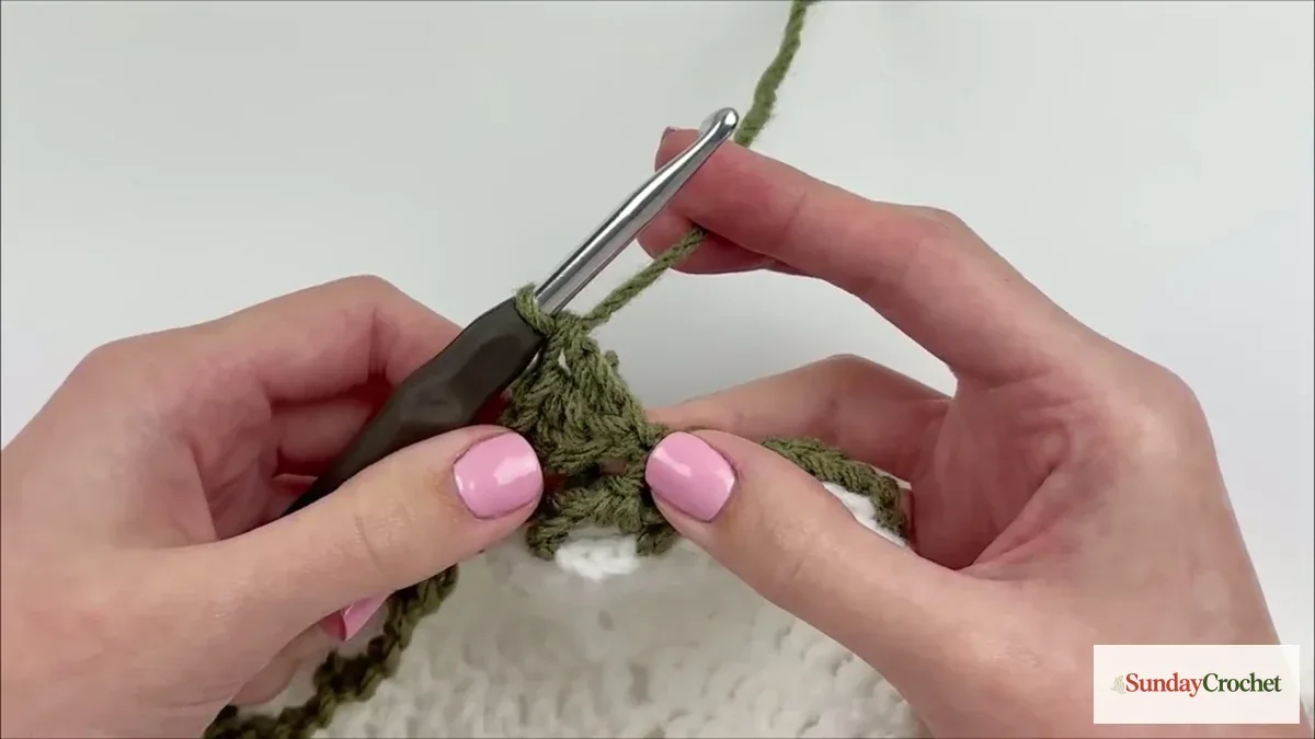 Left-handed crocheter forms a sage-green leaf along the edge of the ivory blanket