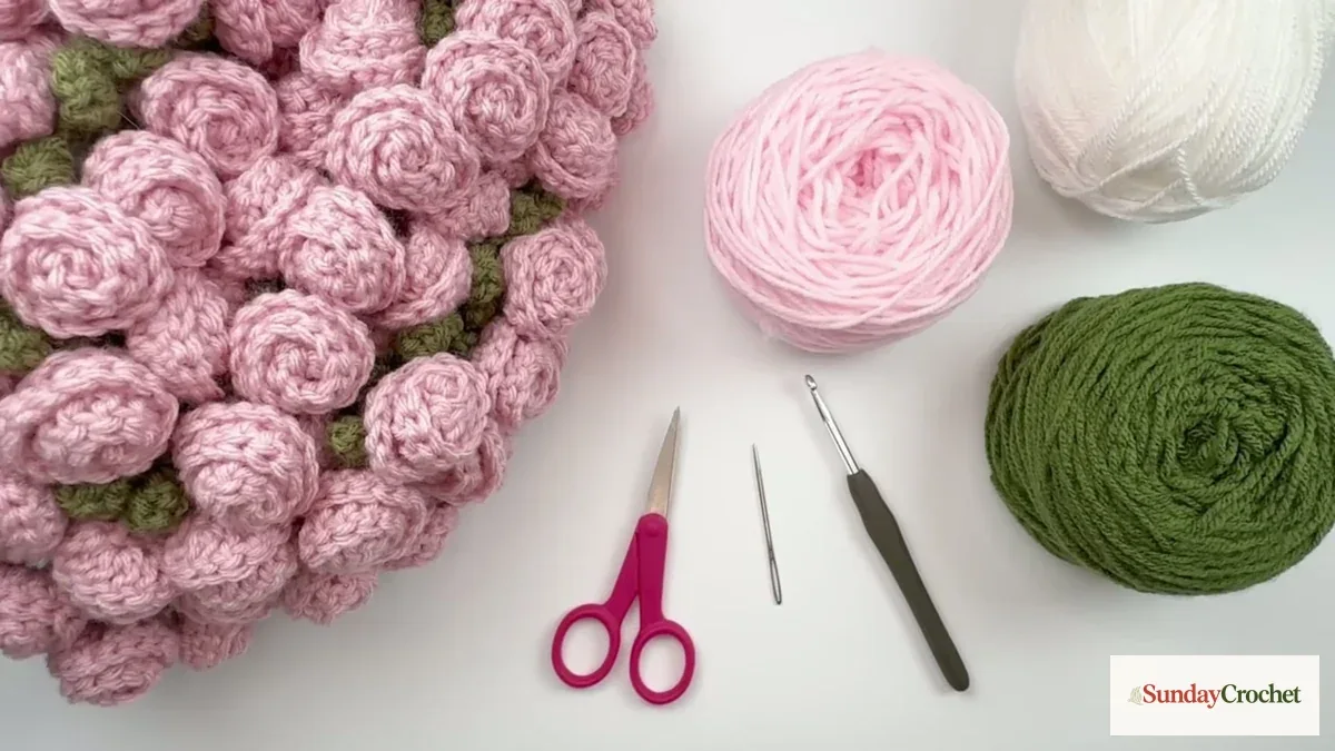 Ivory, pink, and sage yarn with scissors, a crochet hook, and a tapestry needle beside the rose blanket