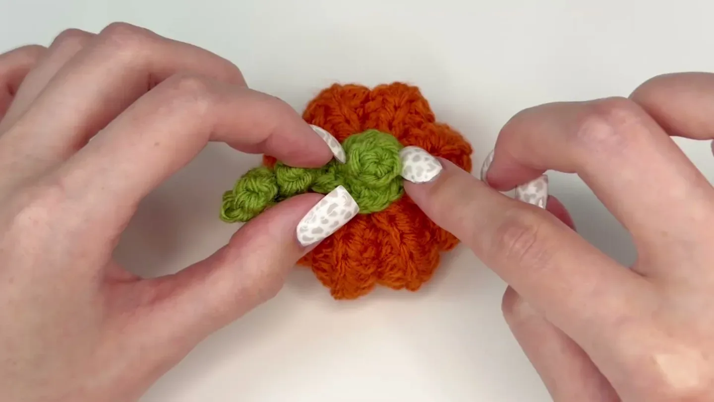 Hands position the finished green stem and vine on top of the orange crochet pumpkin