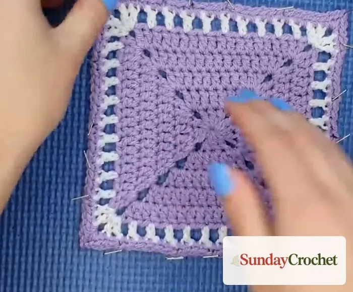 Hands checking a blocked lavender and white crochet square while it remains pinned flat on a blue mat