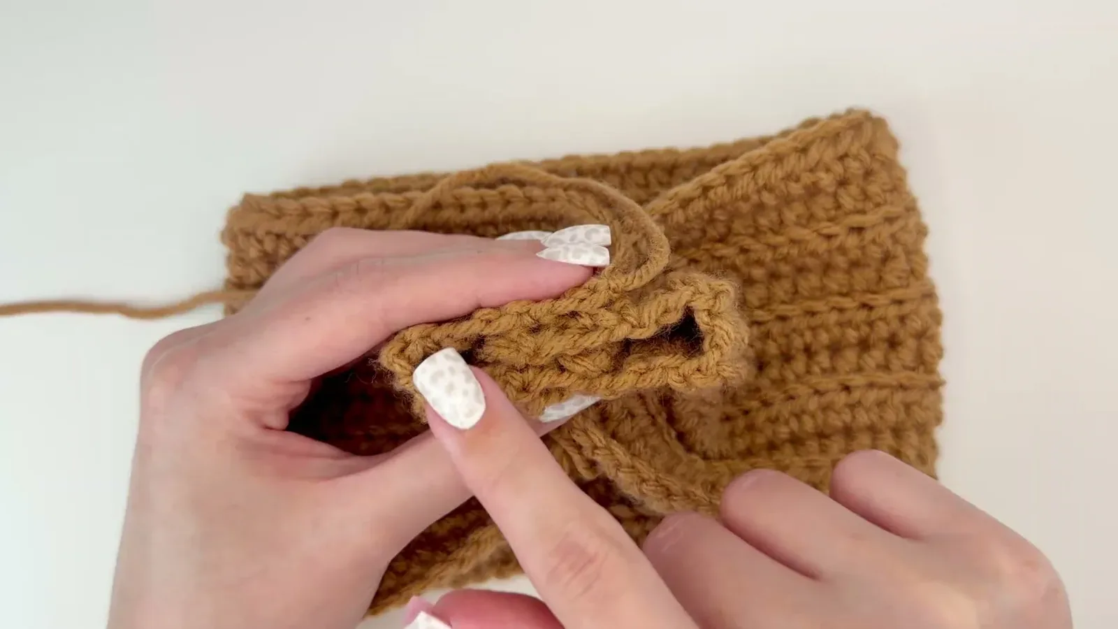 Two folded ends of a caramel crocheted headband being layered to form the center twist