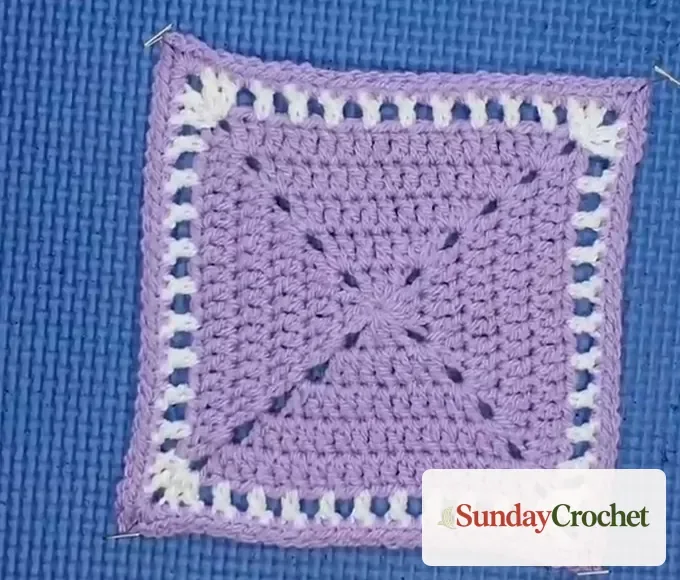 Lavender granny square pinned at its four corners on a blue foam mat