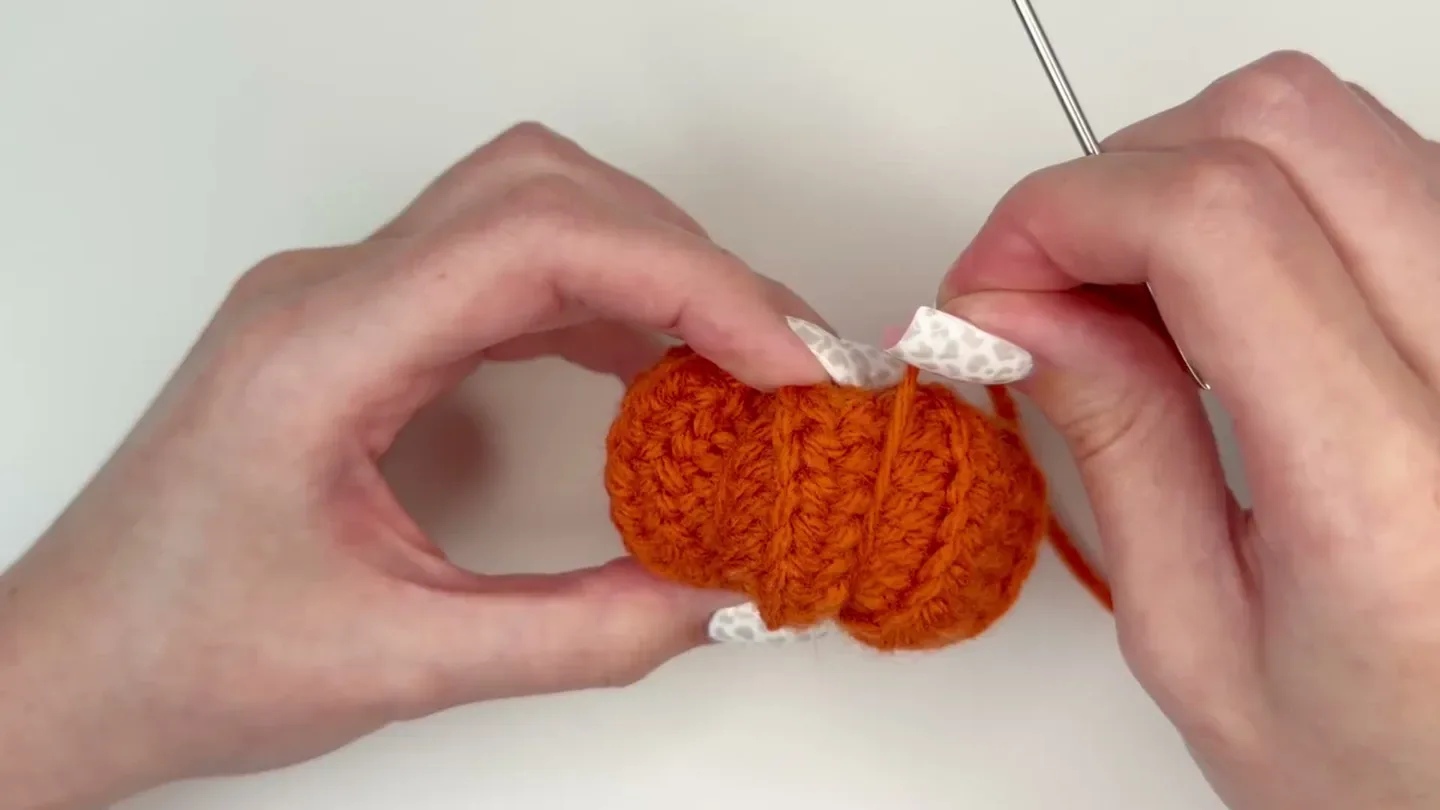 A yarn tail is pulled between the top and bottom of the stuffed orange pumpkin to shape a rib