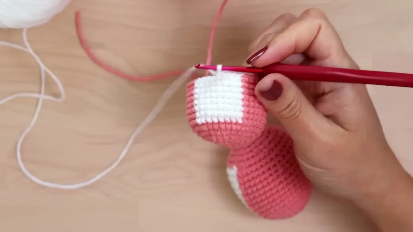A crochet hook lifts a white loop at the color boundary on a pink amigurumi piece.