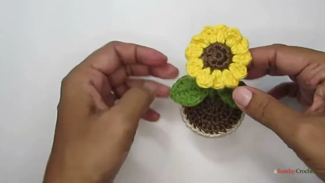 Hands hold a crocheted sunflower above a cream pot of brown soil, with one green leaf visible.