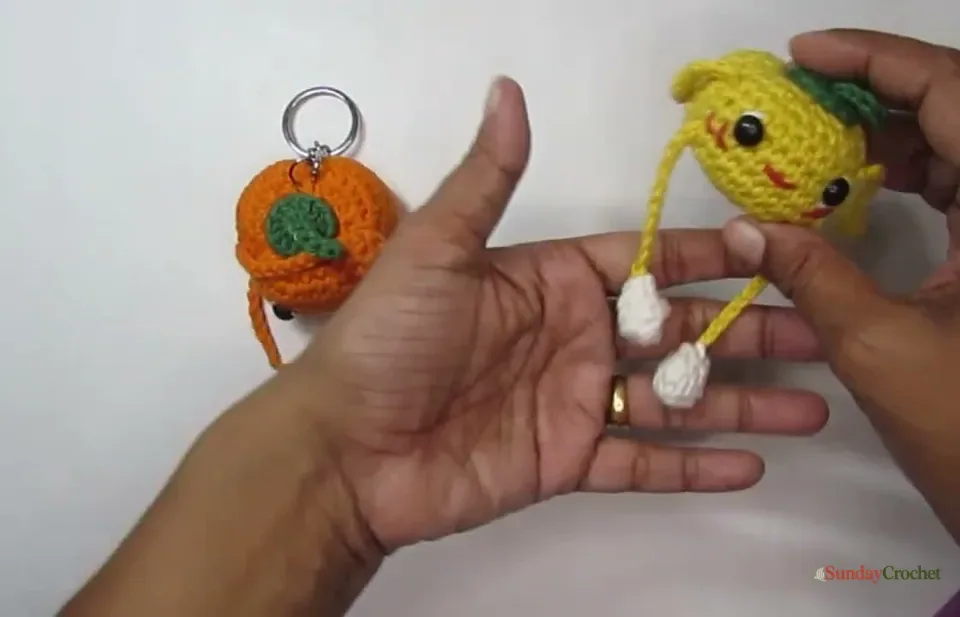 A finished yellow crochet pumpkin with a green stem, long dangling legs, tiny white shoes, and a smiling face