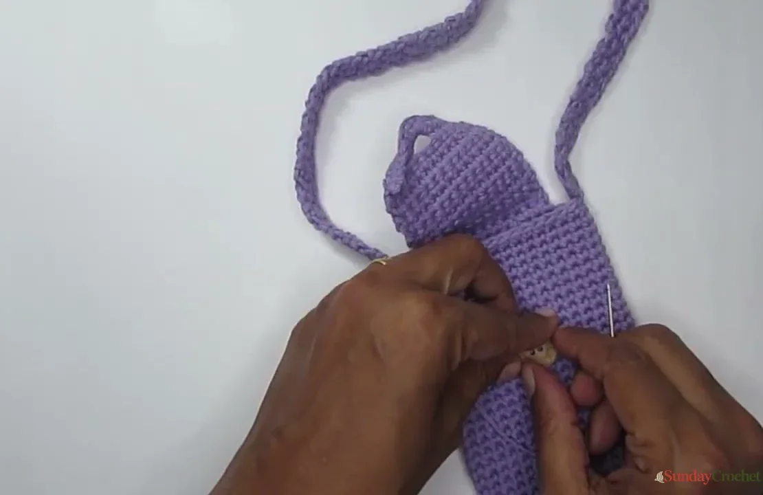 The crochet pouch is held open while a wooden button is positioned and sewn to the front
