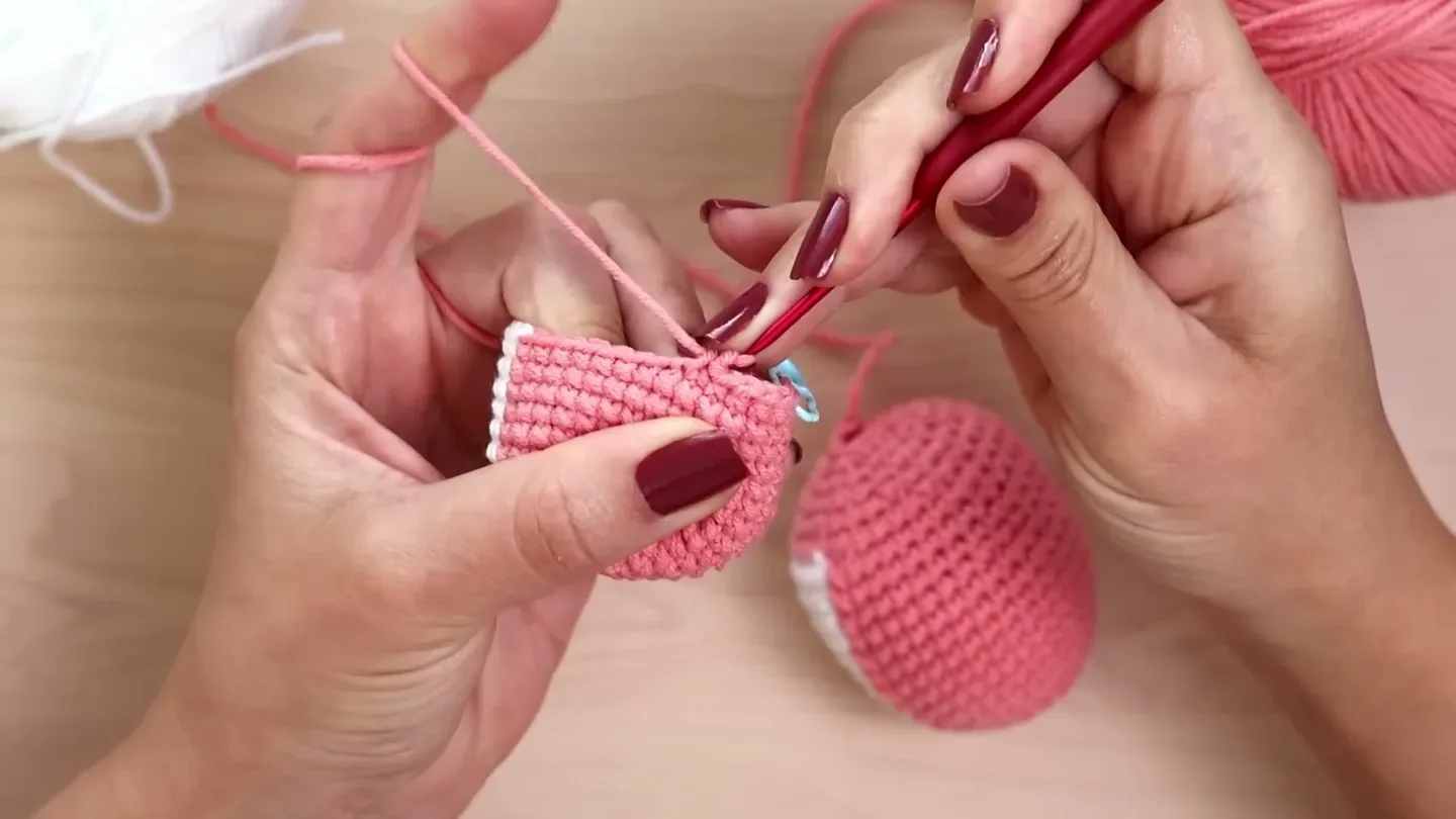 Hands draw the working loop across the top of a pink amigurumi piece while making a single crochet.