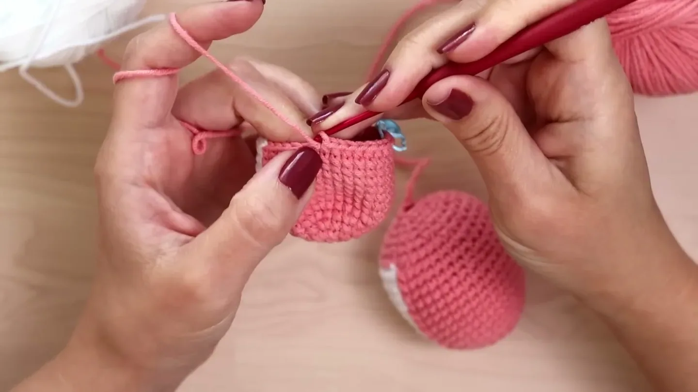 A hook and working yarn lift the loop upward from a single crochet in a pink amigurumi piece.