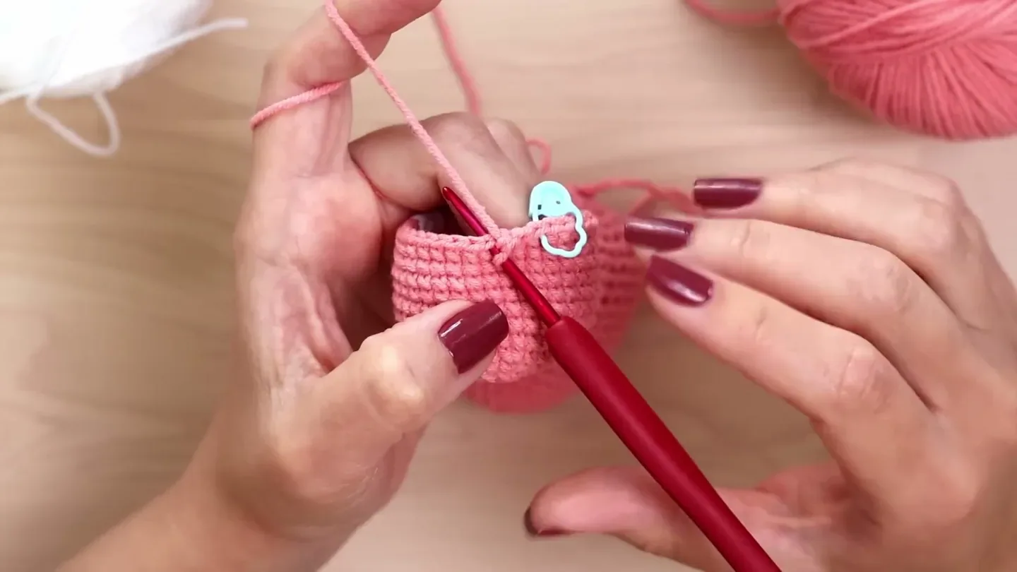 A red crochet hook works a yarn-under single crochet into the top edge of a pink amigurumi tube beside a stitch marker.