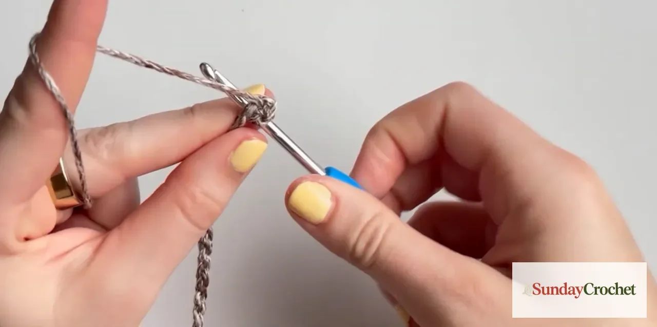 Hands crochet a five-double-crochet cluster in taupe yarn