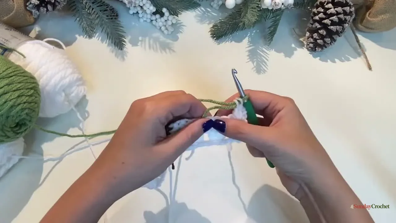 Hands add green yarn to the white granny-stitch fabric to begin the tree motif